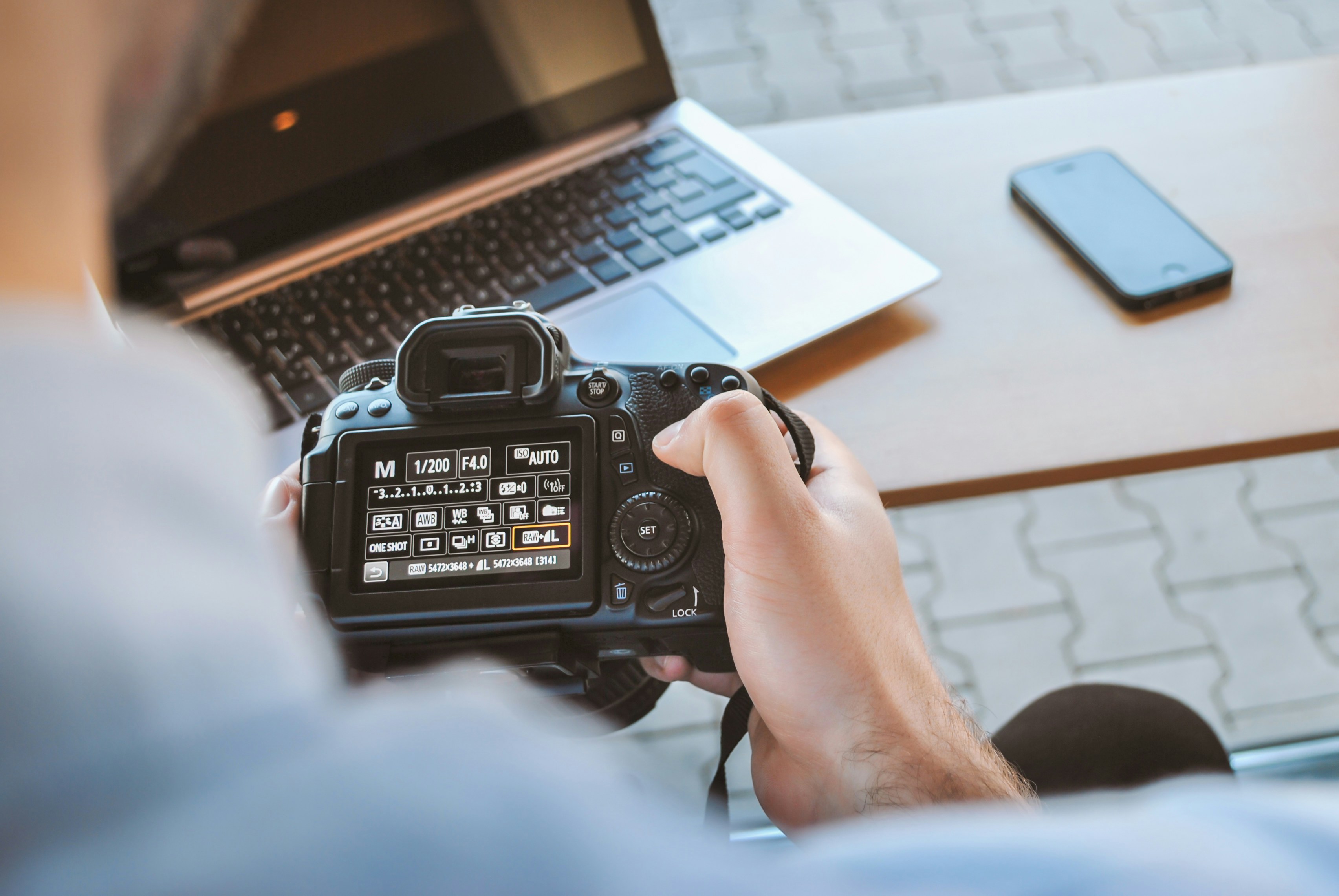 Fotógrafo ajusta configurações da câmera ao lado do notebook e celular, preparação técnica essencial para usar sites para vender fotos.