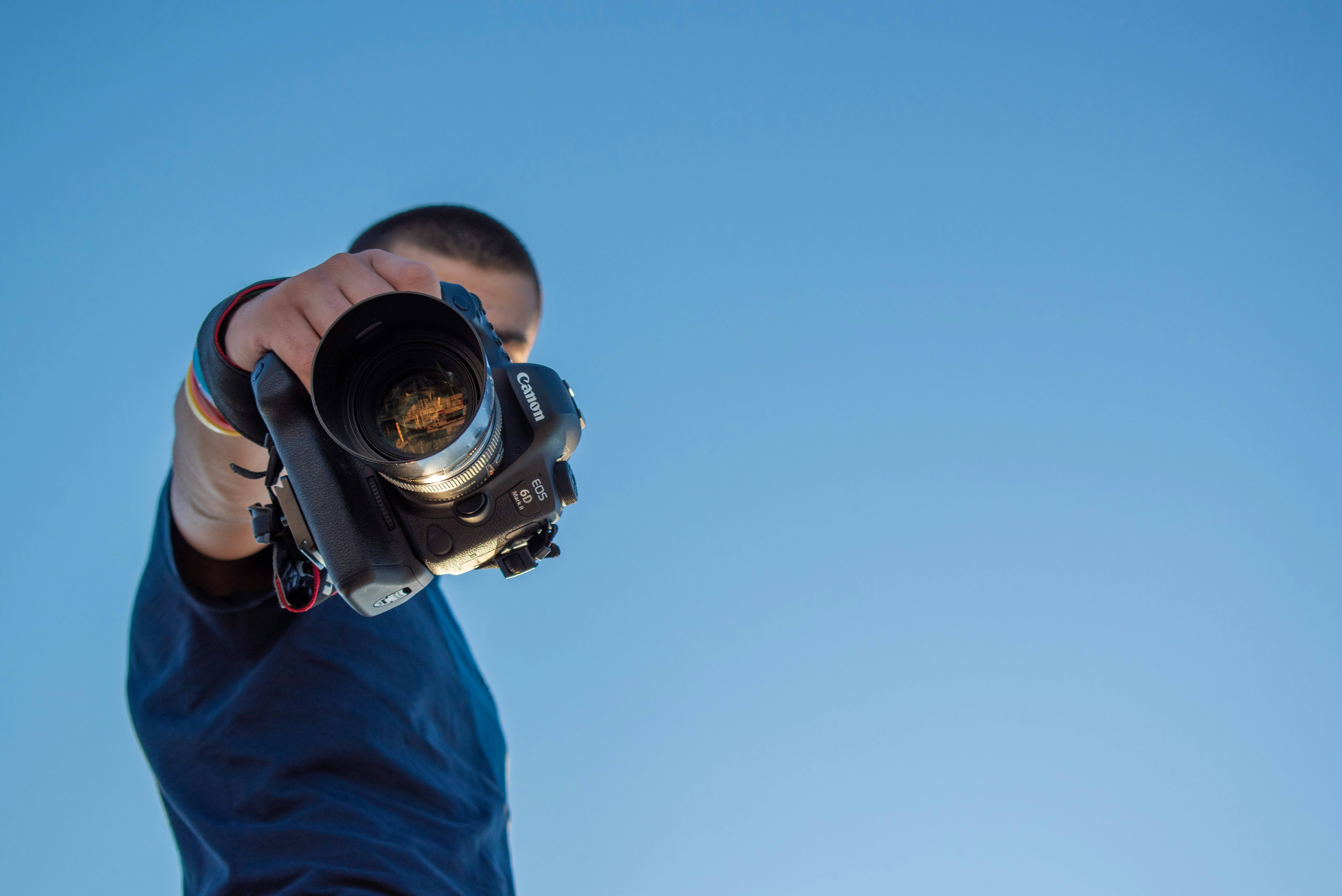 Homem estende o braço apontando uma câmera DSLR para baixo contra o céu azul, visto de ângulo baixo. Exemplo de enquadramento na fotografia.